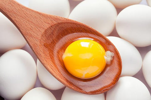 An egg yolk on a wooden spoon with some shelled white eggs in the background
